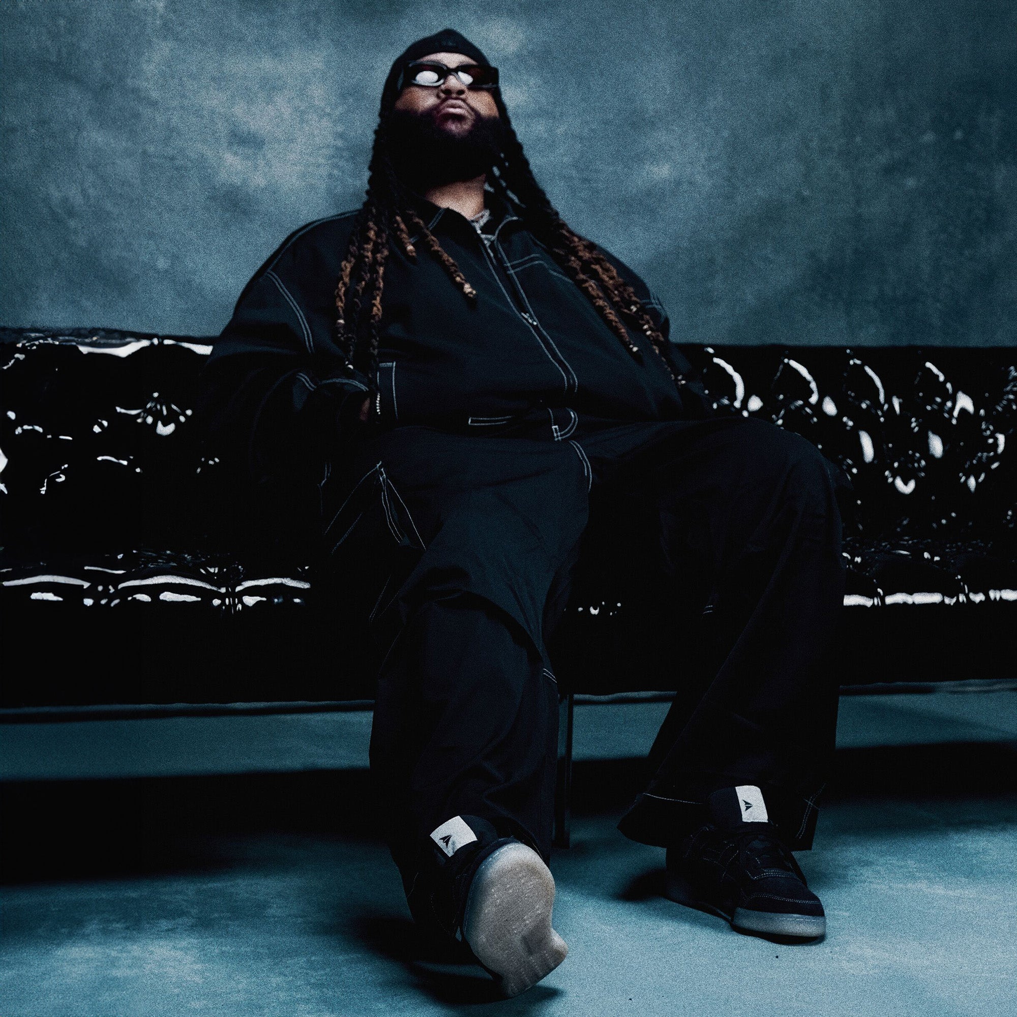 Person wearing a black outfit sitting on a black couch against a dark background
