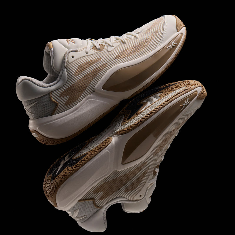 The Reebok Engine A 26 Basketball Shoes feature beige and tan uppers, textured soles, white laces, and mesh detailing for enhanced stability, elegantly reflected on a glossy black surface.