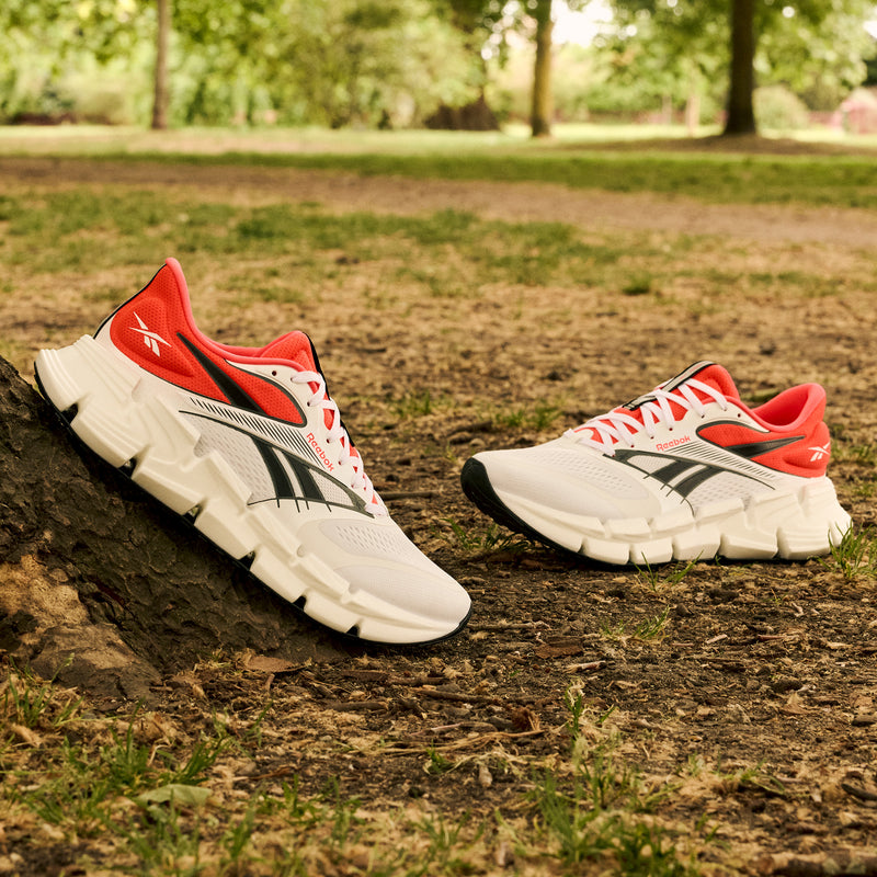 A pair of Reebok Men's FloatZig 2 Running Shoes in white with black and red accents rest on park grass, one leaning against a tree. Superfloat+ cushioning offers comfort, and the shoes stand out amid the greenery.