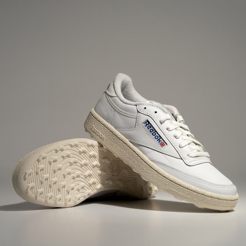 A pair of Reebok Men's Club C Golf Shoes in white with cream spikeless outsoles, one upright and the other propped to display the textured sole and classic logo, set against a plain gray background.
