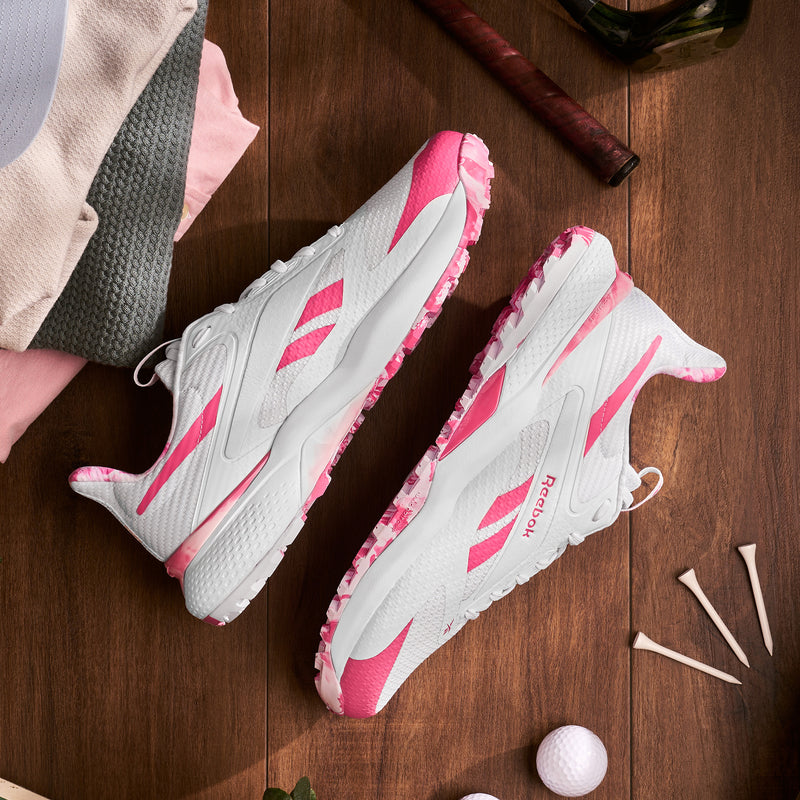 A pair of Reebok Nano Golf Shoes, white with pink accents and Floatride foam for comfort, are displayed on a wooden surface beside golf tees, a golf ball, and folded clothes.