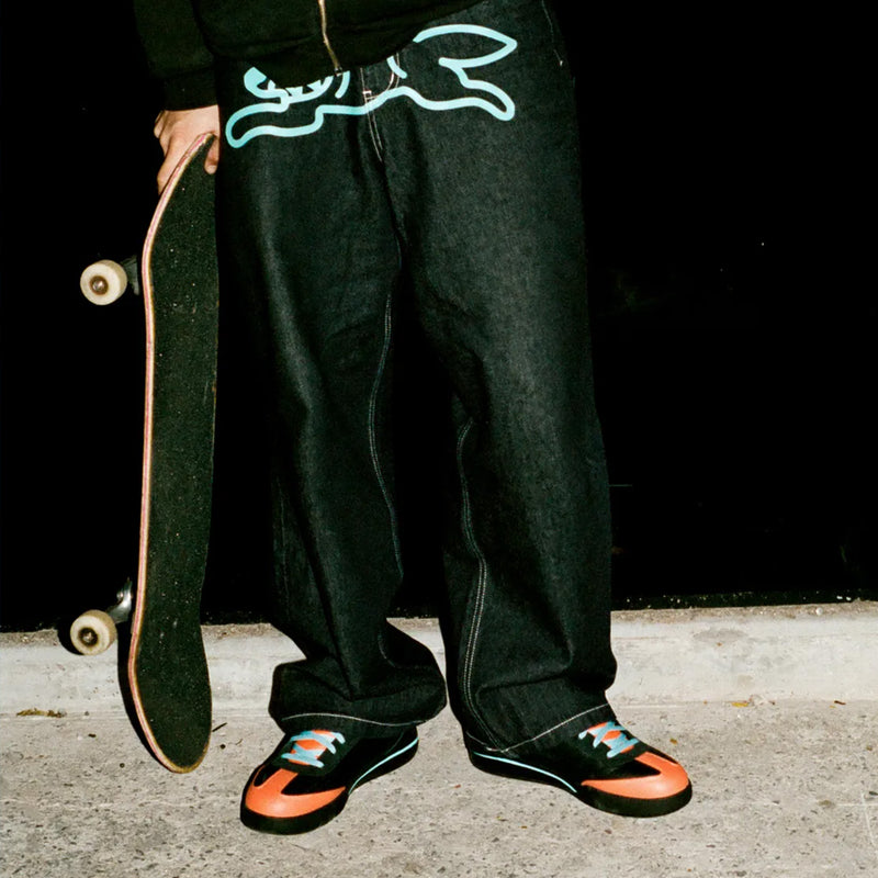A person wearing baggy dark jeans and Reebok x Icecream Board Flip Shoes with black uppers, orange toe caps, and light blue laces, holding a skateboard.