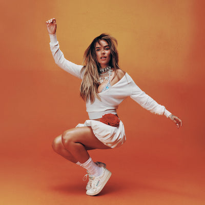 A woman with long hair poses mid-squat against an orange background, looking confidently at the camera. She wears a white cropped sweater, short skirt, layered necklaces, a red belt bag, and completes her look with Reebok Women’s Club C 85 Vintage Shoes.