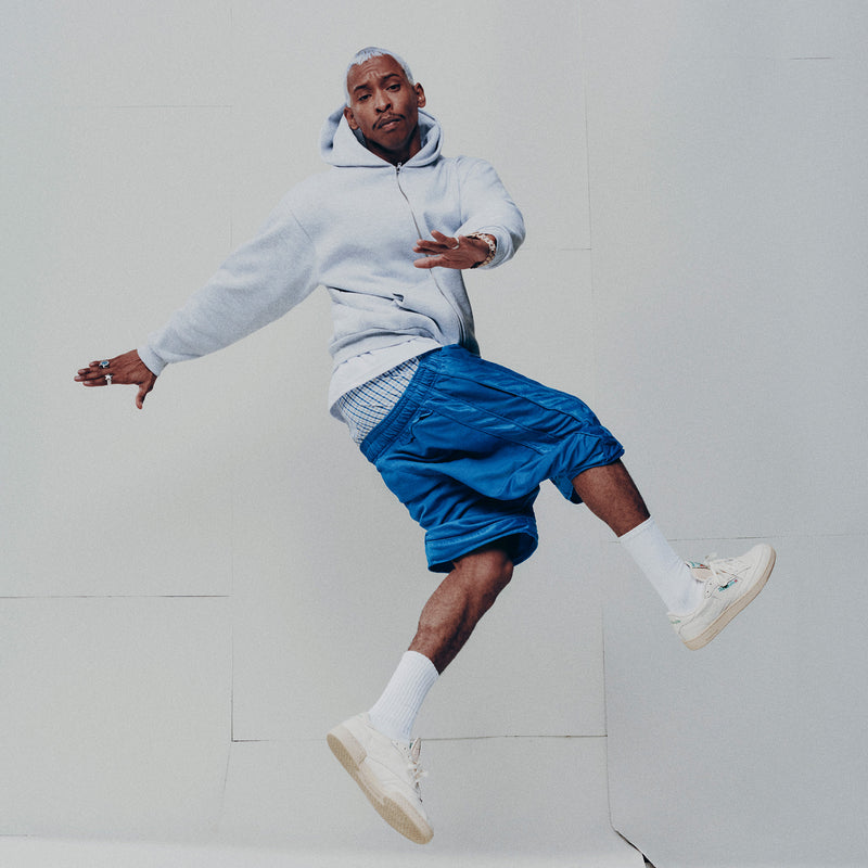 A man in a gray hoodie and blue basketball shorts jumps in the air against a plain light background, wearing Reebok Club C 85 Vintage Shoes featuring a leather upper and lightweight cushioning.