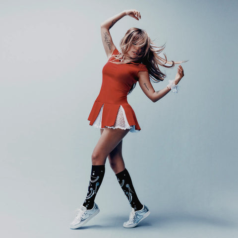 A woman in a red dress and black knee-high socks with white patterns dances, her hair and dress swaying. She wears Reebok Women’s Club C 85 Shoes, which feature a metallic leather upper and an EVA midsole—perfectly complementing her tattooed arms as she poses against a plain light blue background.