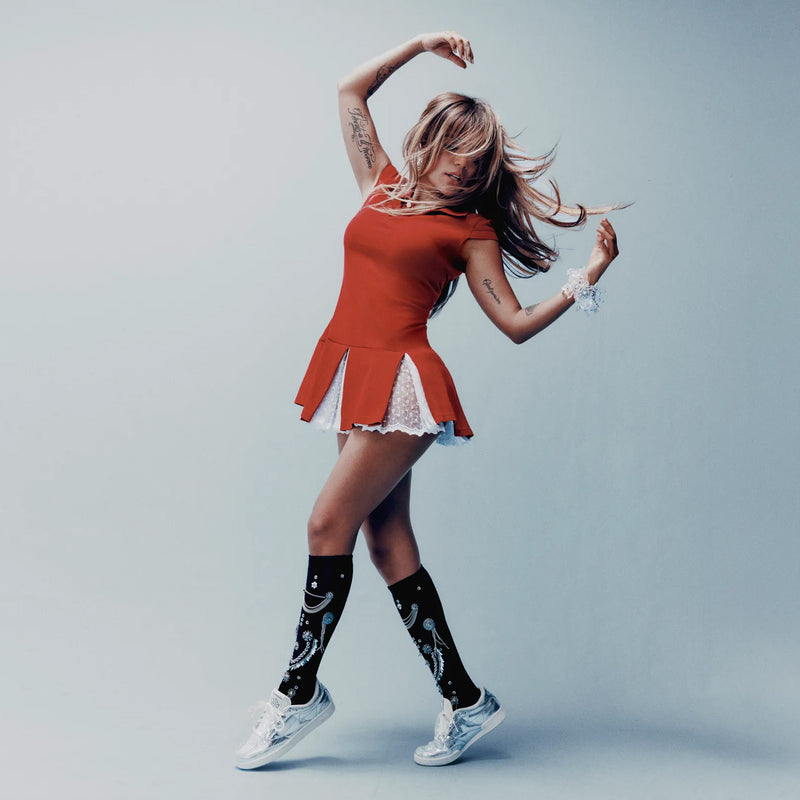 A woman in a red dress and black knee-high socks with white patterns dances, her hair and dress swaying. She wears Reebok Women’s Club C 85 Shoes, which feature a metallic leather upper and an EVA midsole—perfectly complementing her tattooed arms as she poses against a plain light blue background.