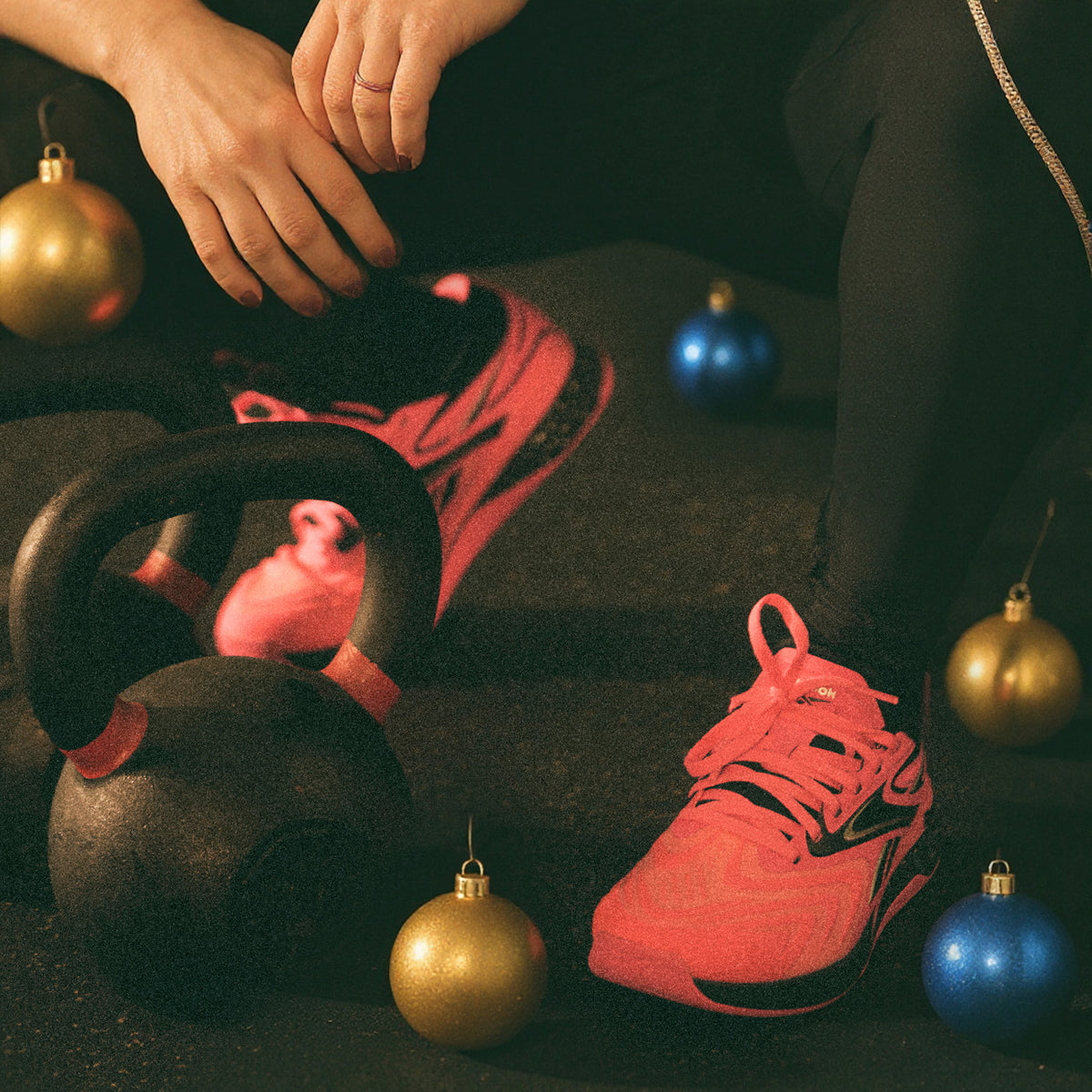 A pair of bright coral-pink running shoes styled on a dark floor with a kettlebell, a person's hands and leg visible, and several gold and blue Christmas ornaments.