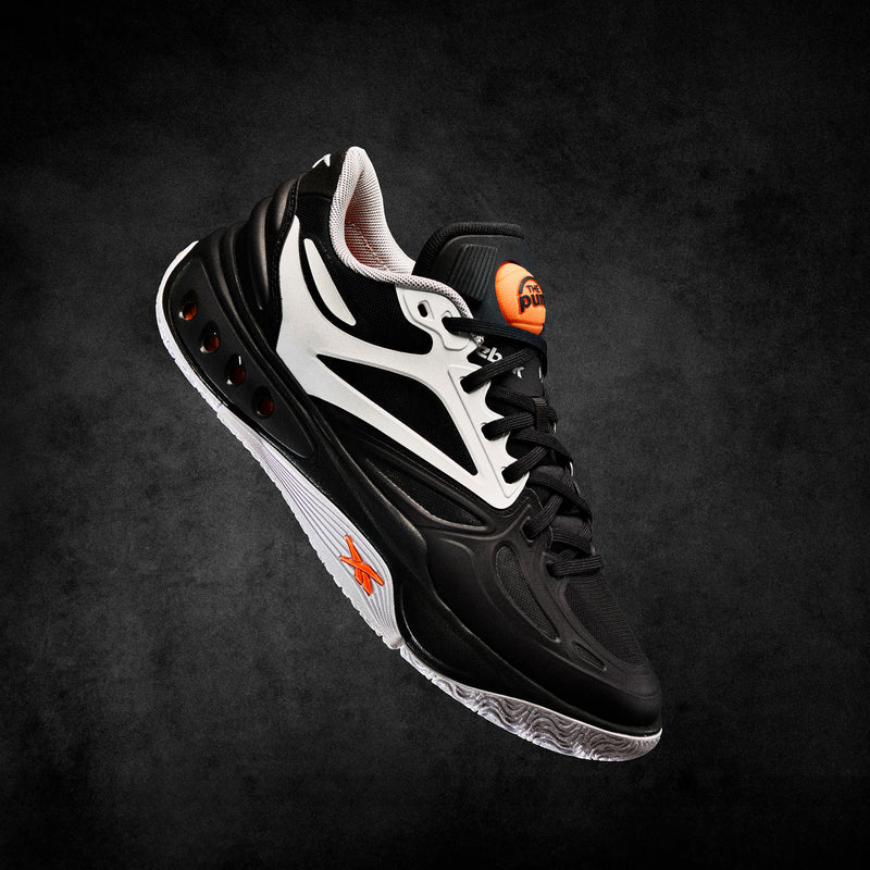 A black and white Reebok Engine A Pump Shoe with striking orange accents floats against a dark textured background, highlighting its side profile, innovative Pump technology, and intricate design details.