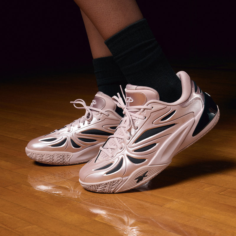 A person stands on a polished gym floor wearing Reebok Angel Reese 1 Basketball Shoes—shiny, futuristic light pink performance sneakers with black details—and black socks.