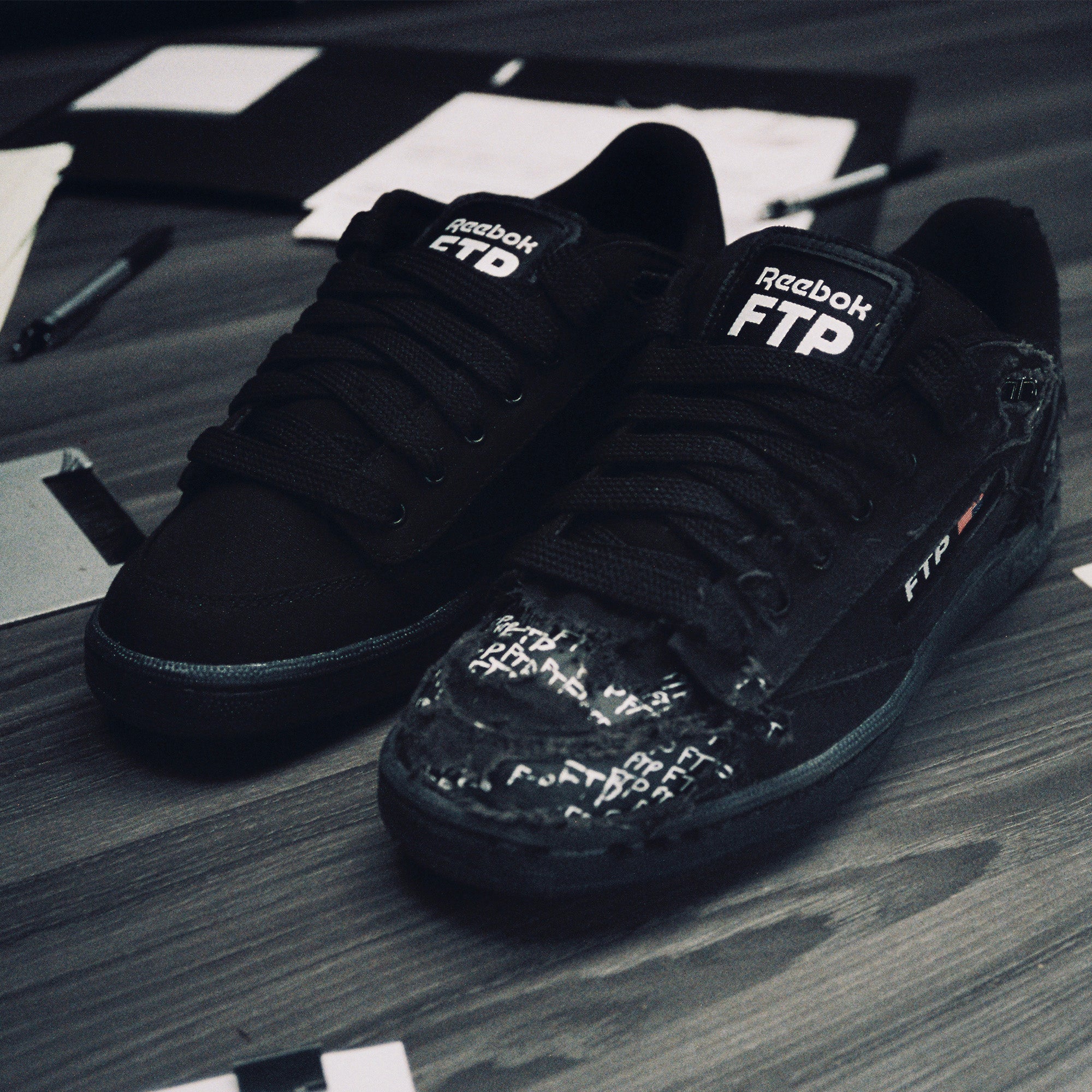 Black Reebok FTP sneakers on a wooden surface with scattered papers and pens.