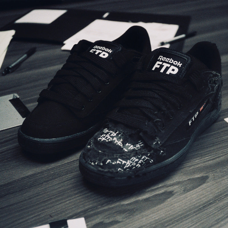 Black Reebok FTP sneakers on a wooden surface with scattered papers and pens.