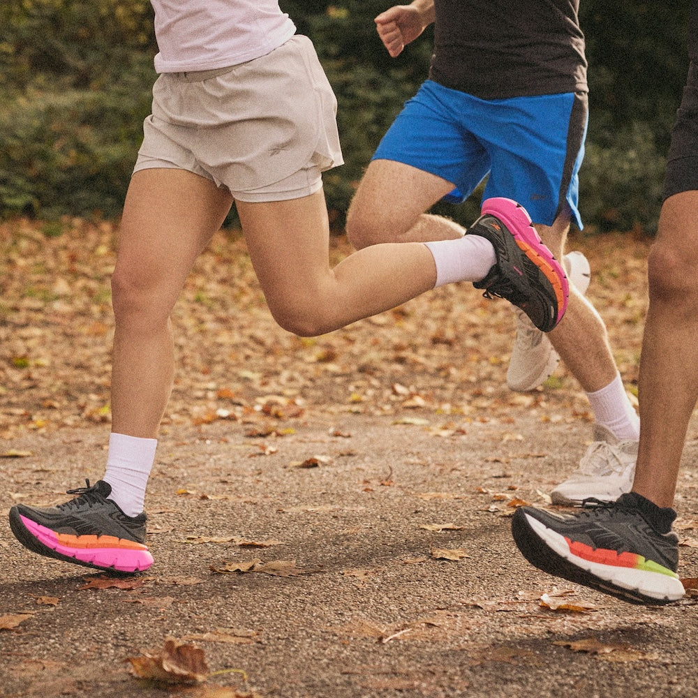 Three people jogging outdoors on a leaf-covered path, wearing athletic shorts, t-shirts, and running shoes. Only their legs and lower bodies are visible in the image. Trees and greenery are in the background.