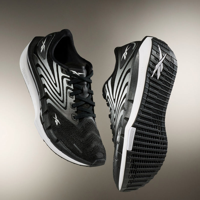 A pair of Reebok Women's FloatZig Tread Running Shoes in black and white is displayed from the side and bottom, showcasing the patterned sole, breathable mesh upper, sleek design with white accents, and Reebok branding—ideal for treadmill workouts.