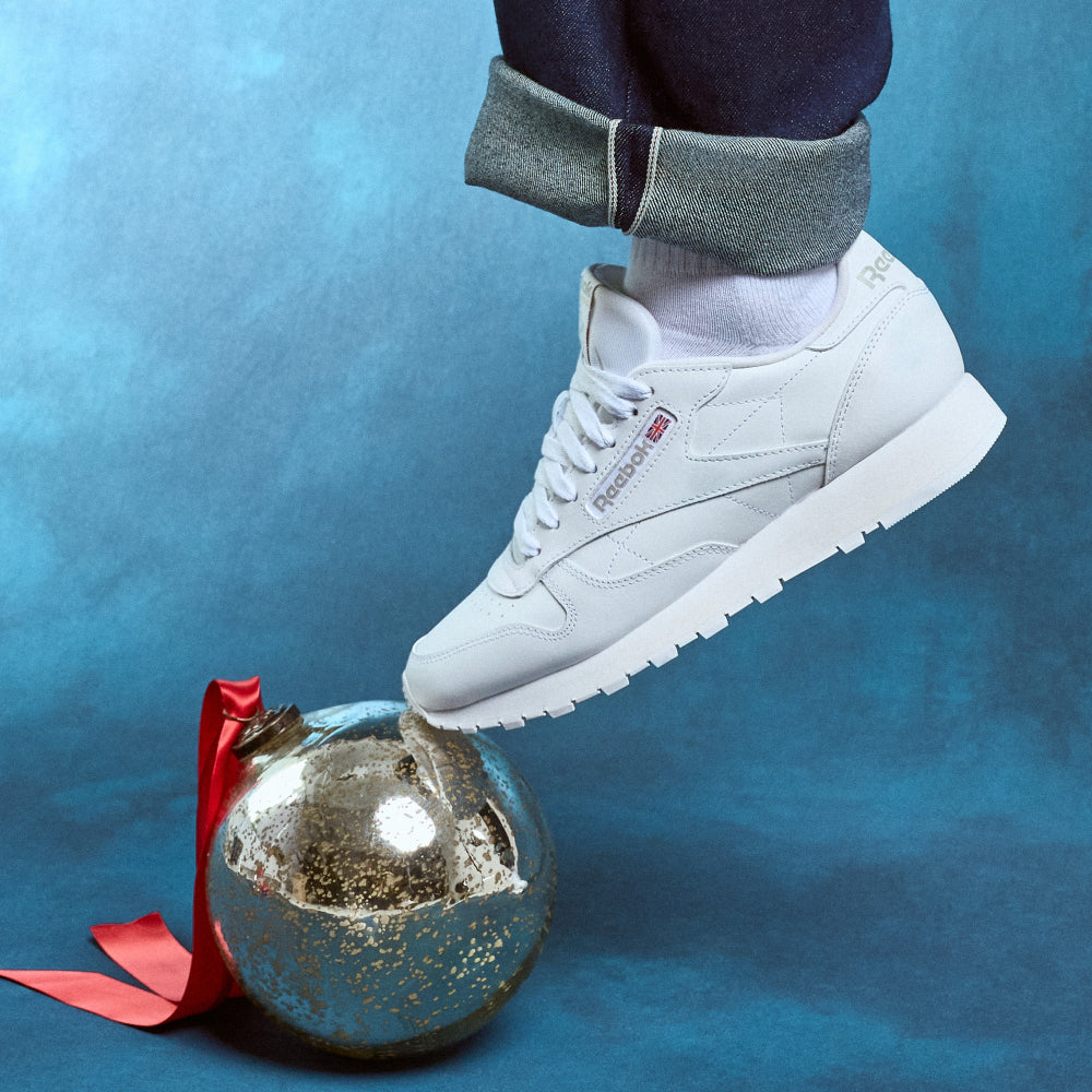 White sneaker stepping onto a reflective silver ornament with a red ribbon against a blue background