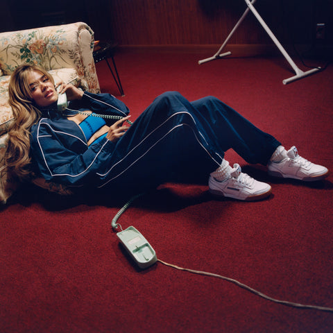 A woman in a blue tracksuit lounges on a red carpet by a floral sofa, chatting on a green corded phone. She wears Reebok Women's Workout Plus Shoes, channeling classic '80s fitness style as the phone cord coils across the floor.