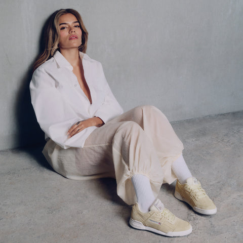 Karol G wearing a white shirt, beige pants, and beige sneakers sitting on the floor against a plain wall.