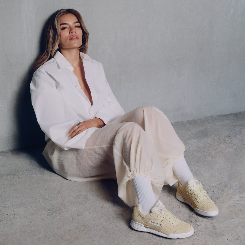Karol G wearing a white shirt, beige pants, and beige sneakers sitting on the floor against a plain wall.