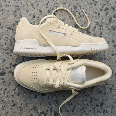 A pair of Women's Workout Plus Shoes by Reebok in beige, with laces, placed on textured gray concrete—one lying flat and the other on its side—evokes classic '80s fitness style.