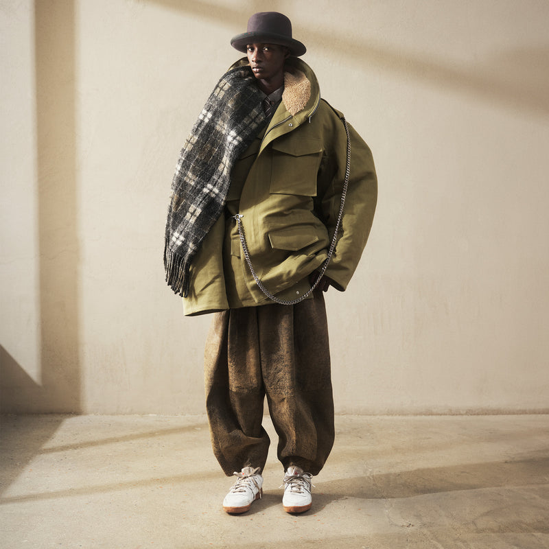 Person wearing a green coat, brown pants, and a plaid scarf against a beige wall.