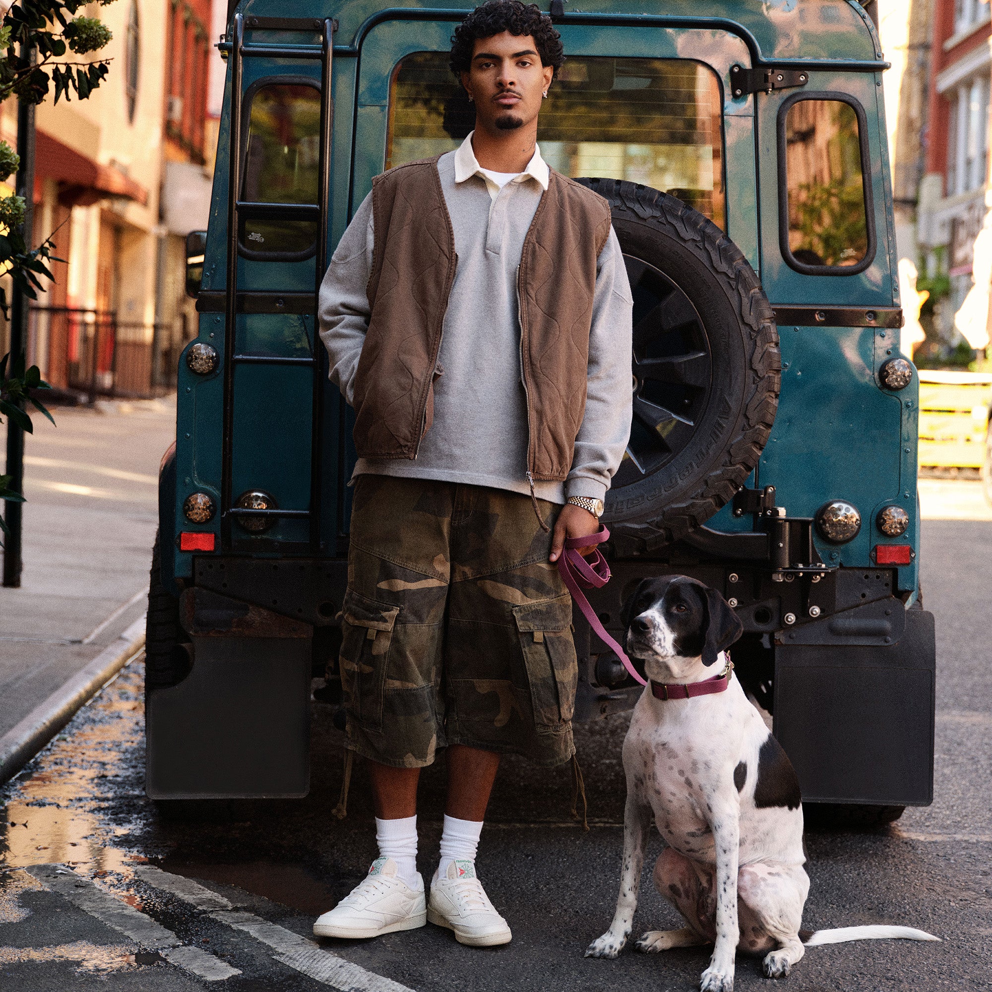 A man wearing Reebok classics shoes standing behind a vehicle on a city street with a dog on a leash.