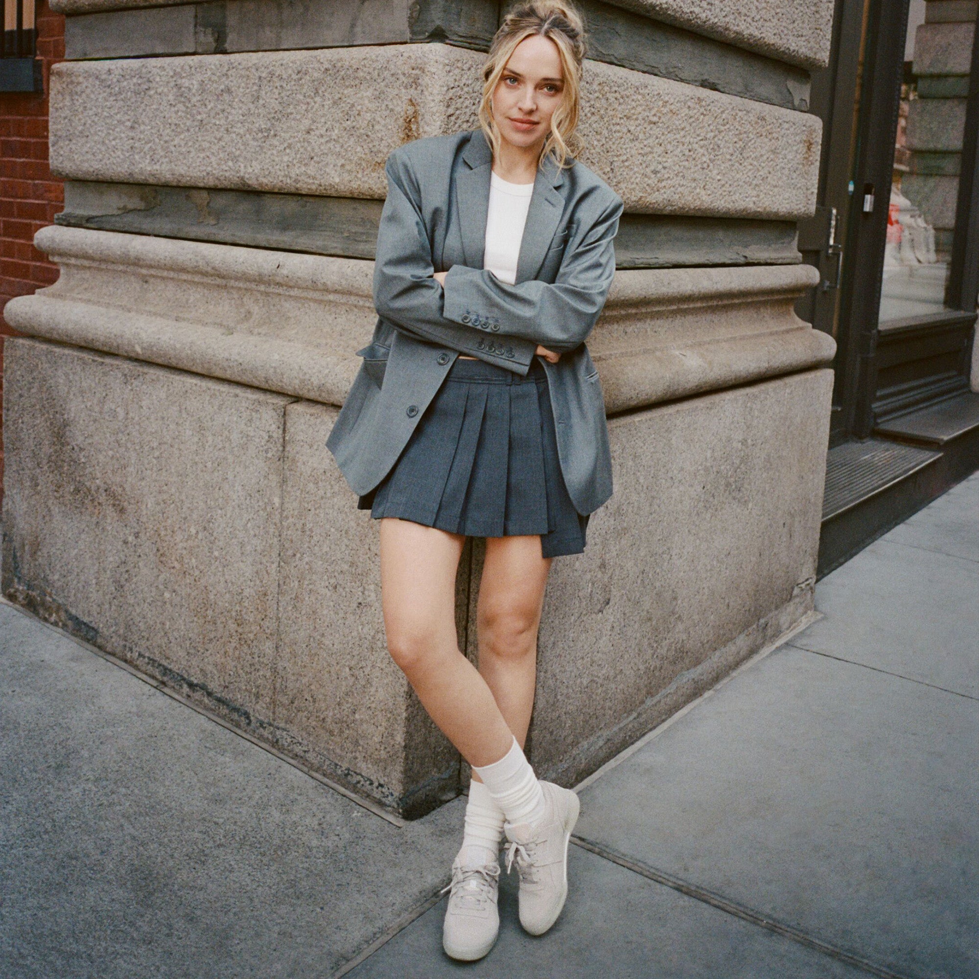 Woman in a gray blazer and pleated skirt standing against a stone wall.