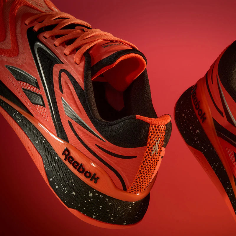 Close-up of two orange Reebok running shoes with black and white accents, seen against a red background, emphasizing the brand logo and modern design details.