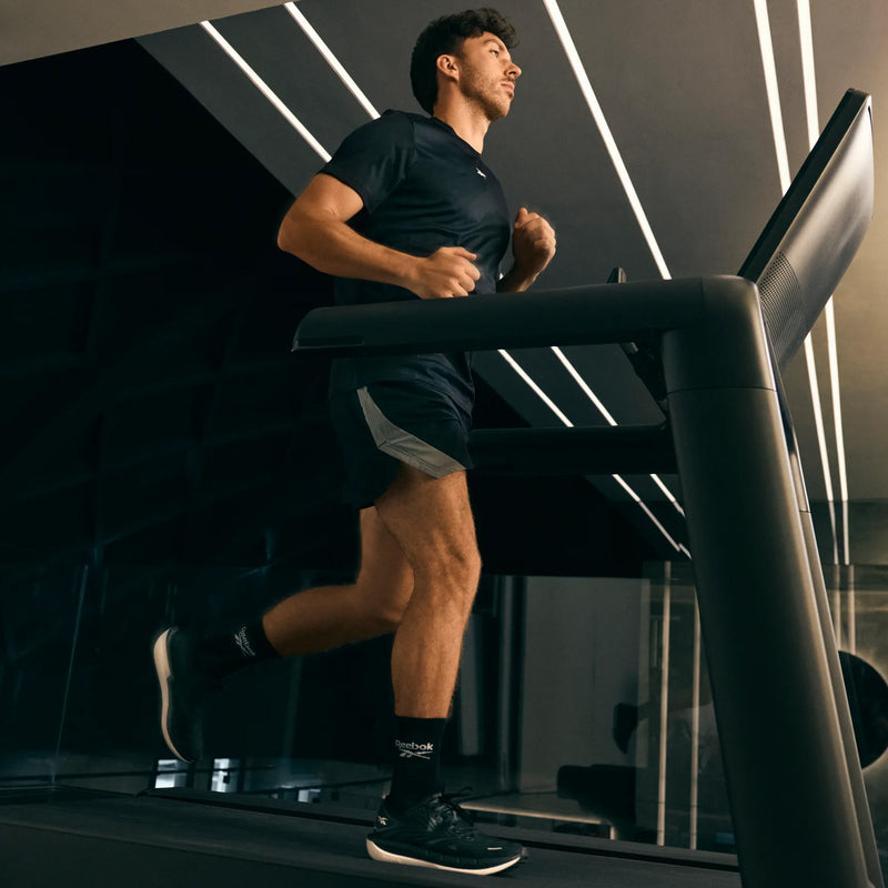 A man in dark athletic wear, sporting Reebok Men’s FloatZig Tread Running Shoes with a cushioned midsole, runs on a treadmill in a modern gym with sleek lighting and reflective surfaces.
