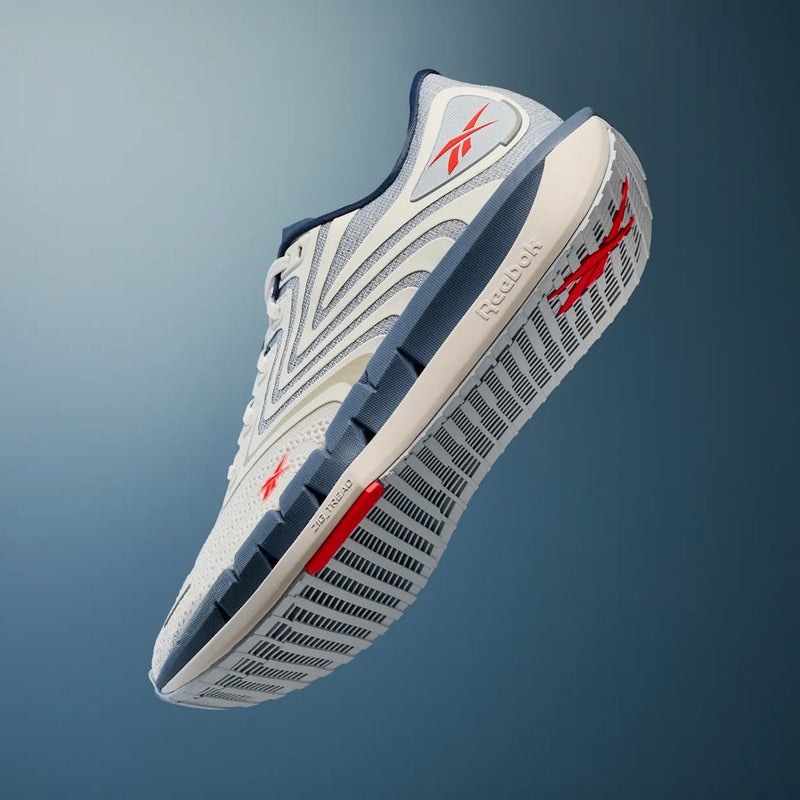 The Reebok Men’s FloatZig Tread Running Shoes in white and blue, featuring bold red accents, are captured from a low angle against a gradient blue background. This shot highlights the cushioned midsole and signature side branding—making them an ideal choice for treadmill workouts or daily runs.