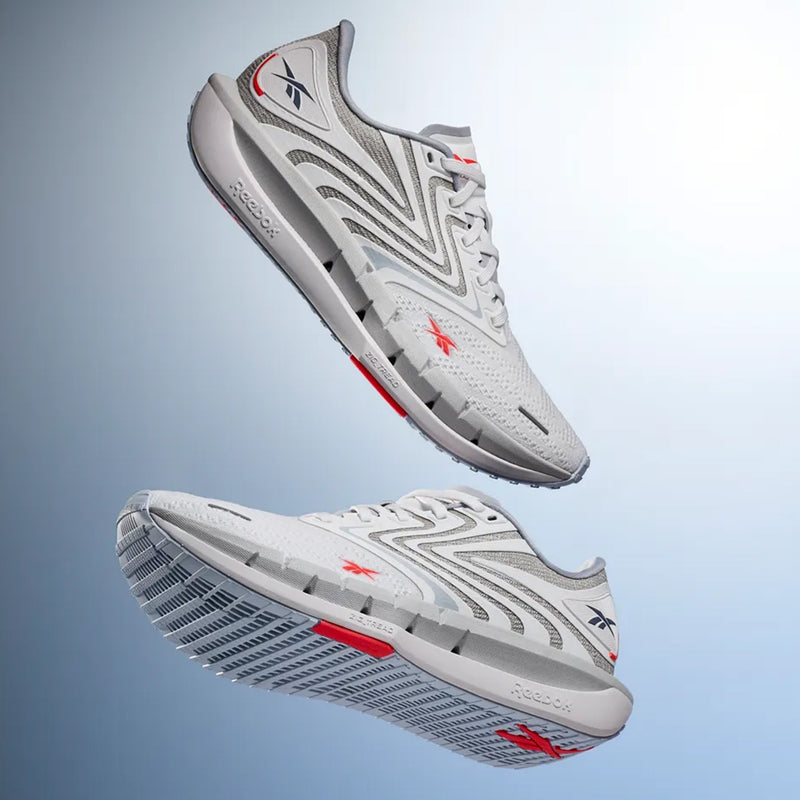 Two Reebok Women’s FloatZig Tread Running Shoes with gray and red accents appear suspended in mid-air against a light blue gradient background, showcasing both the sides and soles. Featuring a sleek design with breathable mesh, they are an ideal choice for treadmill workouts or any active routine.