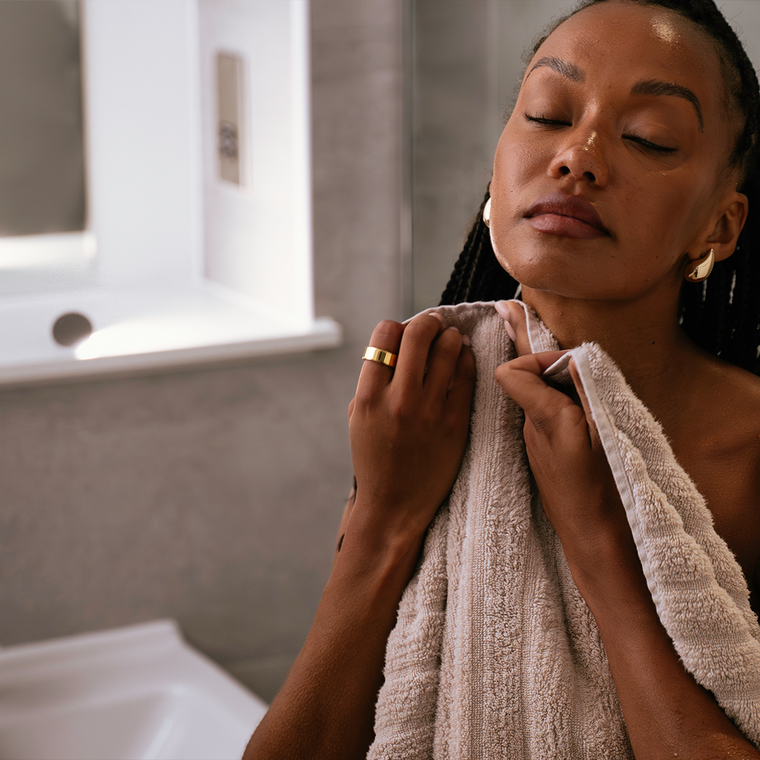 A woman with closed eyes gently dries her neck with a beige towel in a bathroom, wearing a gold ring and earrings, with a serene expression and braided hair.