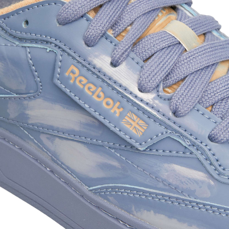 Close-up of a glossy periwinkle blue Reebok Club C LTD low-top sneaker with matching flat laces and a tan tongue, styled as a classic tennis shoe.