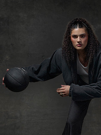 A person with curly hair wearing a black jacket and pants crouches while holding a black basketball, ready to dribble against a dark, textured background.