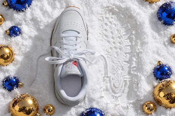 A white Reebok sneaker lies on white snow beside a matching white shoe print, surrounded by blue and gold Christmas ornaments.