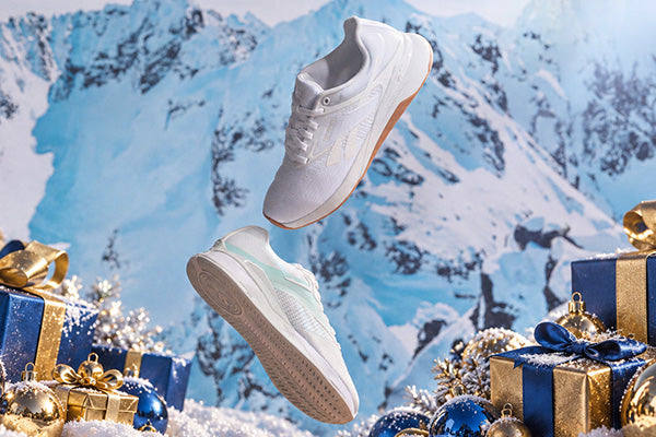 A pair of white athletic sneakers floating in front of a snowy mountain backdrop, surrounded by blue and gold Christmas gift boxes and ornaments.