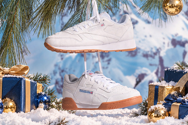 A pair of Reebok Classic Leather sneakers in white with gum brown soles, suspended in a snowy Christmas-themed setting with wrapped gifts, pine branches, and gold ornaments.