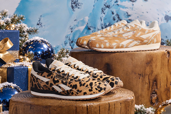 Two pairs of Reebok Classic sneakers styled in a festive winter setting. One pair features a tan and black leopard print with white stripes, and the other features a tan and black dalmatian pattern with white stripes.