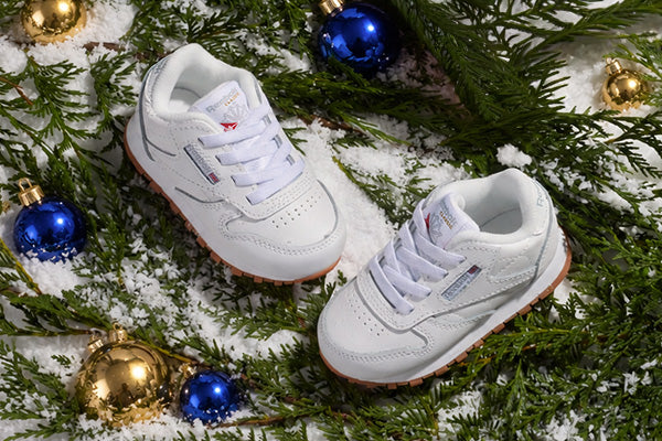 A pair of white Reebok sneakers styled in a festive Christmas setting, surrounded by pine branches, snow, and gold and blue ornaments.