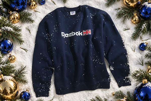 A navy blue Reebok crewneck sweatshirt with a white Reebok logo and Union Jack graphic on the chest, styled flat-lay on white faux snow with pine branches and blue and gold Christmas ornaments.