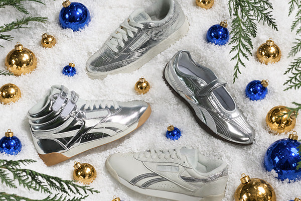 Four Reebok sneakers in metallic silver, black, and white colorways arranged on a white snowy surface decorated with blue and gold Christmas ornaments and pine branches. Reebok branding is visible on the side of the shoes.