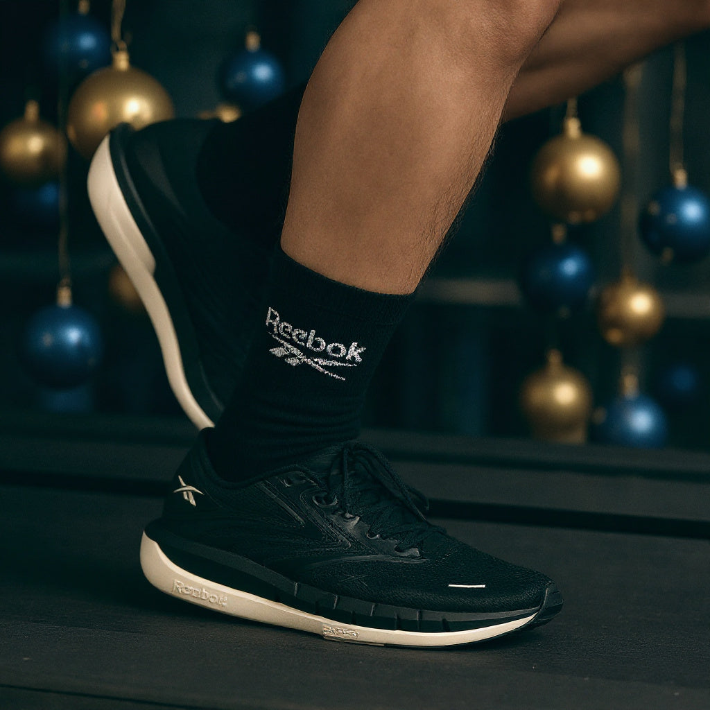 A person wearing black Reebok athletic socks and black Reebok running shoes, with the Reebok logo clearly visible on the sock and side of the shoe. The background is dark with blurred blue and gold hanging ornaments.