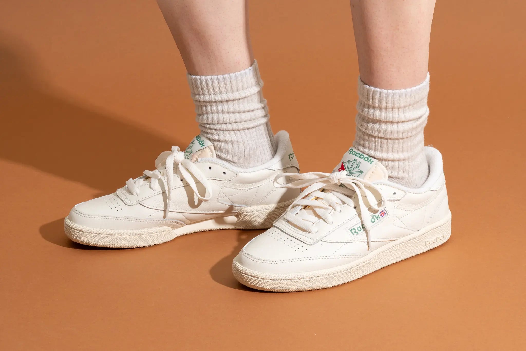 A person wearing white socks and off-white Reebok sneakers stands on a light brown surface. The sneakers feature green and red Reebok logos on the tongues and sides.