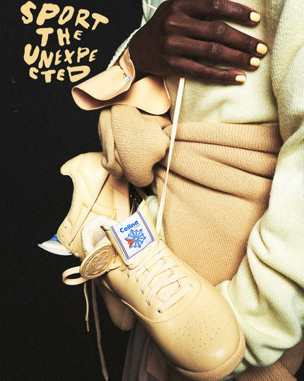 Model holding a butter yellow Celine x Reebok Freestyle Lo sneaker, styled in tonal cream and camel knitwear, with “Sport the Unexpected” handwritten in the background.