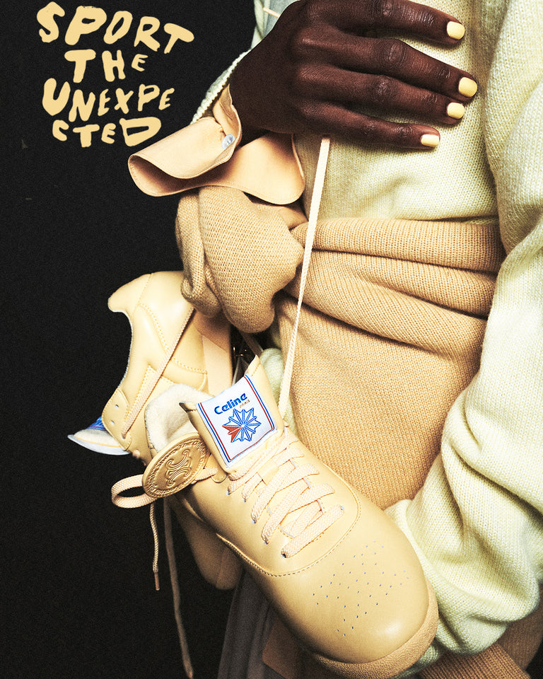 Model holding a butter yellow Celine x Reebok Freestyle Lo sneaker, styled in tonal cream and camel knitwear, with “Sport the Unexpected” handwritten in the background.