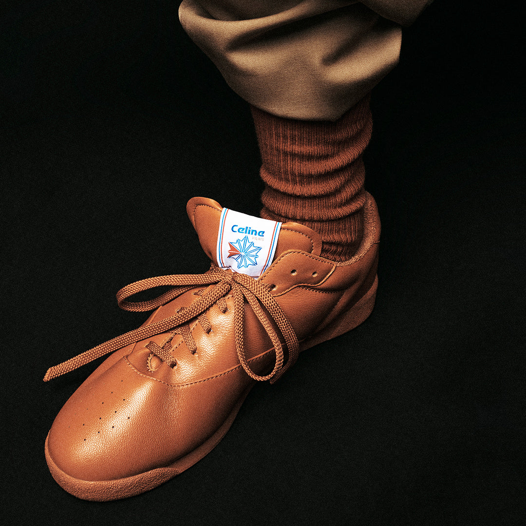 Close-up of a brown CELINE Reebok sneaker with tonal laces and socks against a black background.