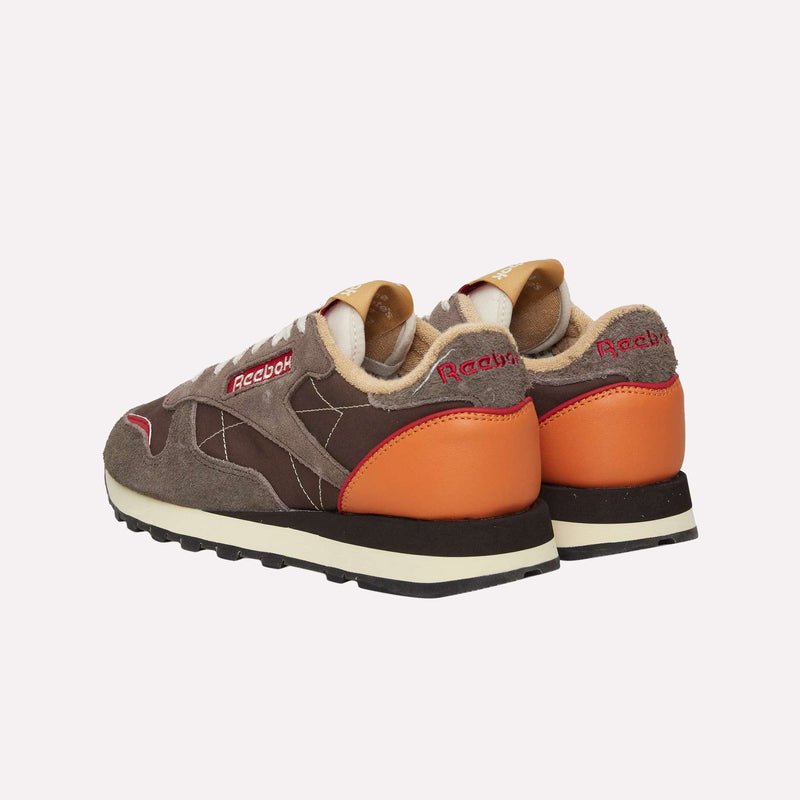 A pair of Reebok Classic Leather Premium LTD Shoes in brown, featuring orange heel accents, beige lining, and white soles, are positioned facing away at an angle on a plain light background, highlighting their classic silhouette inspired by timeless retro sneakers.