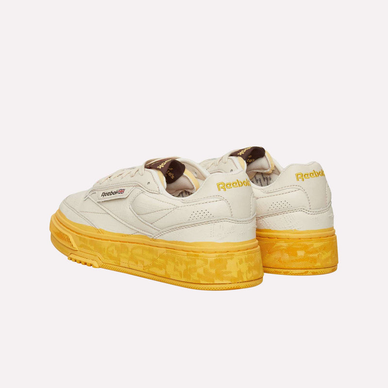 Two cream-colored Reebok Club C LTD Shoes with yellow soles and yellow Reebok branding on the heel, displayed against a plain white background for a timeless lifestyle classic tennis shoe look.