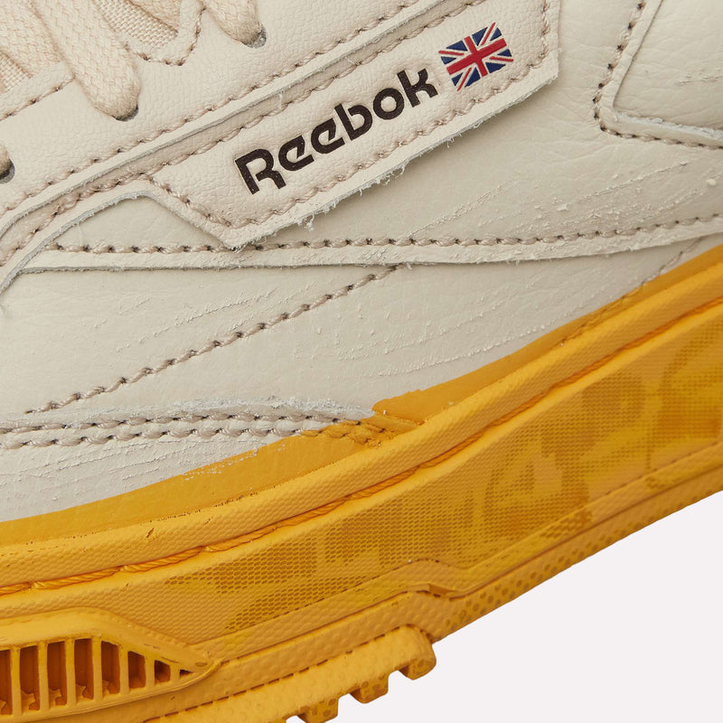 A close-up of the Reebok Club C LTD Shoes in white, featuring a yellow sole, detailed stitching, and a small Union Jack flag beside the Reebok logo—this modern lifestyle classic draws inspiration from heritage tennis footwear.