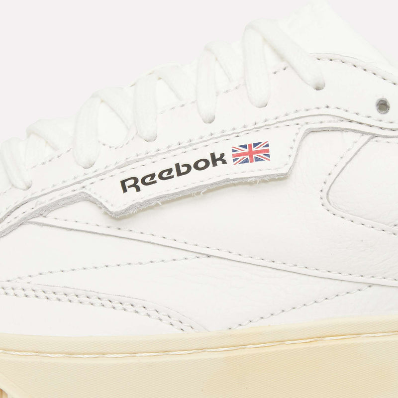 Close-up of a white Reebok Club C LTD Shoe, highlighting the Reebok logo and small British flag on the side. This iconic tennis shoe features white laces and detailed stitching atop a cream-colored sole, making it a true lifestyle classic.