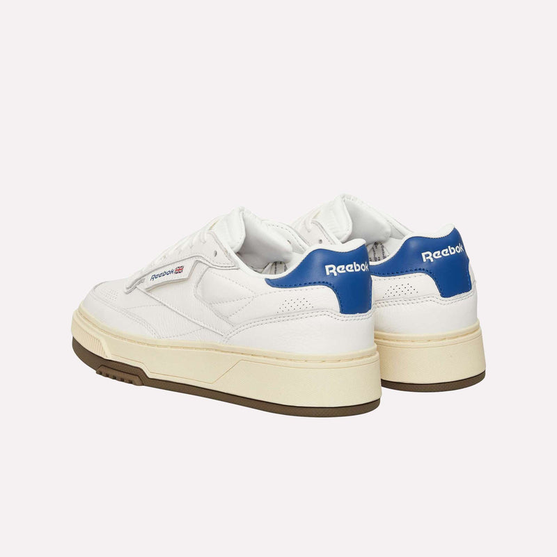 A pair of Reebok Club C LTD Shoes in white, featuring blue heel accents, cream-colored midsoles, and gum outsoles. These lifestyle classics display the Reebok branding on the side and heel, merging tennis-inspired heritage with enduring style.