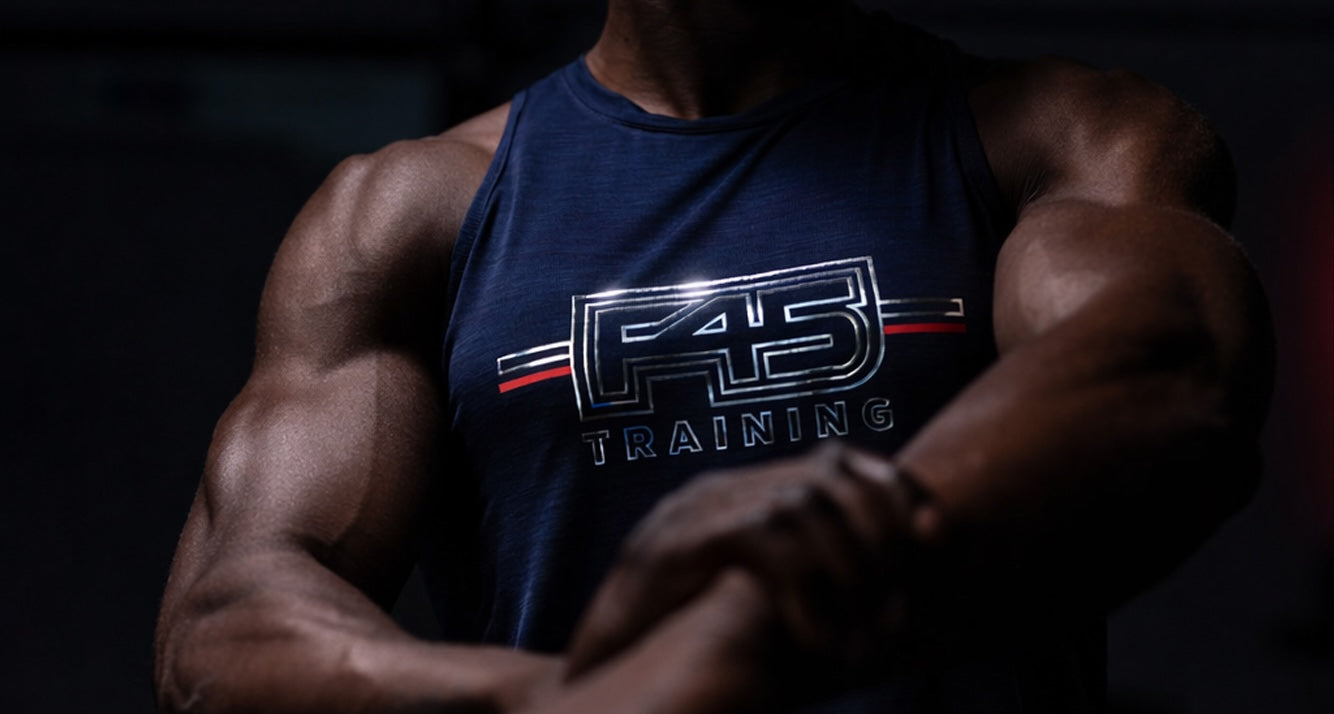 A muscular person wearing a sleeveless navy shirt with F45 Training written on it, shown from shoulders to waist, flexing their arm in a dimly lit setting.